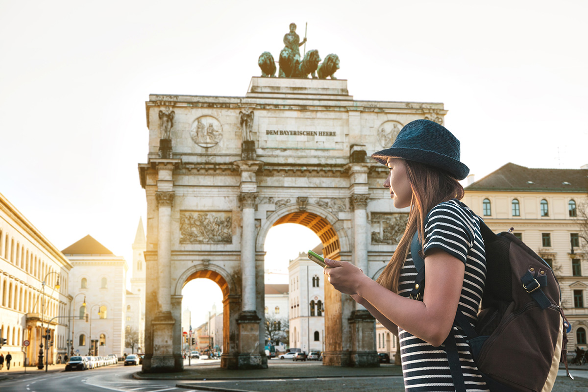 A person in a striped shirt and hat holds a smartphone while standing in front of the Siegestor, a triumphal arch captured beautifully, as if from a German textbook. The sun casts a warm glow on this iconic landmark and surrounding Munich buildings.