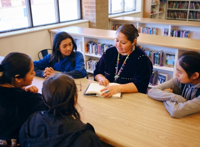 Teacher seated at a table with a group of students. They are listening to the teacher while she reads a book. 