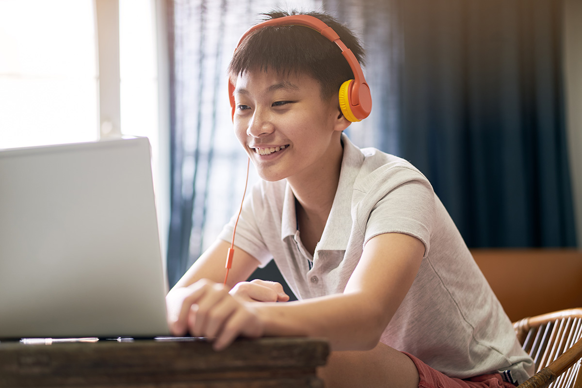 A person wearing orange headphones sits at a desk, smiling while engaging in a listening comprehension activity on their laptop.