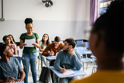 A girl in a classroom standing up with a piece of paper, reading aloud while her classmates look at her. 