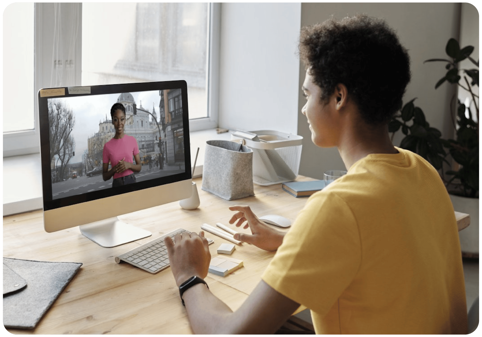 A student wearing a yellow shirt sits at a desk using an Apple computer with ClearTalk, an AI-powered speaking tool. The screen shows an individual in a pink shirt against a city backdrop. The desk is adorned with stationery, a plant, and has the light of a window nearby.