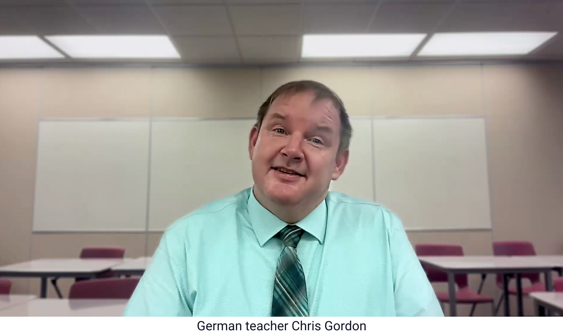 German teacher Chris Gordon, sits in a classroom with whiteboards and rows of desks and chairs behind him.