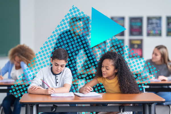 A picture of two students working together in a classroom, with pencils and notebooks. Other students and a teacher are in the background.